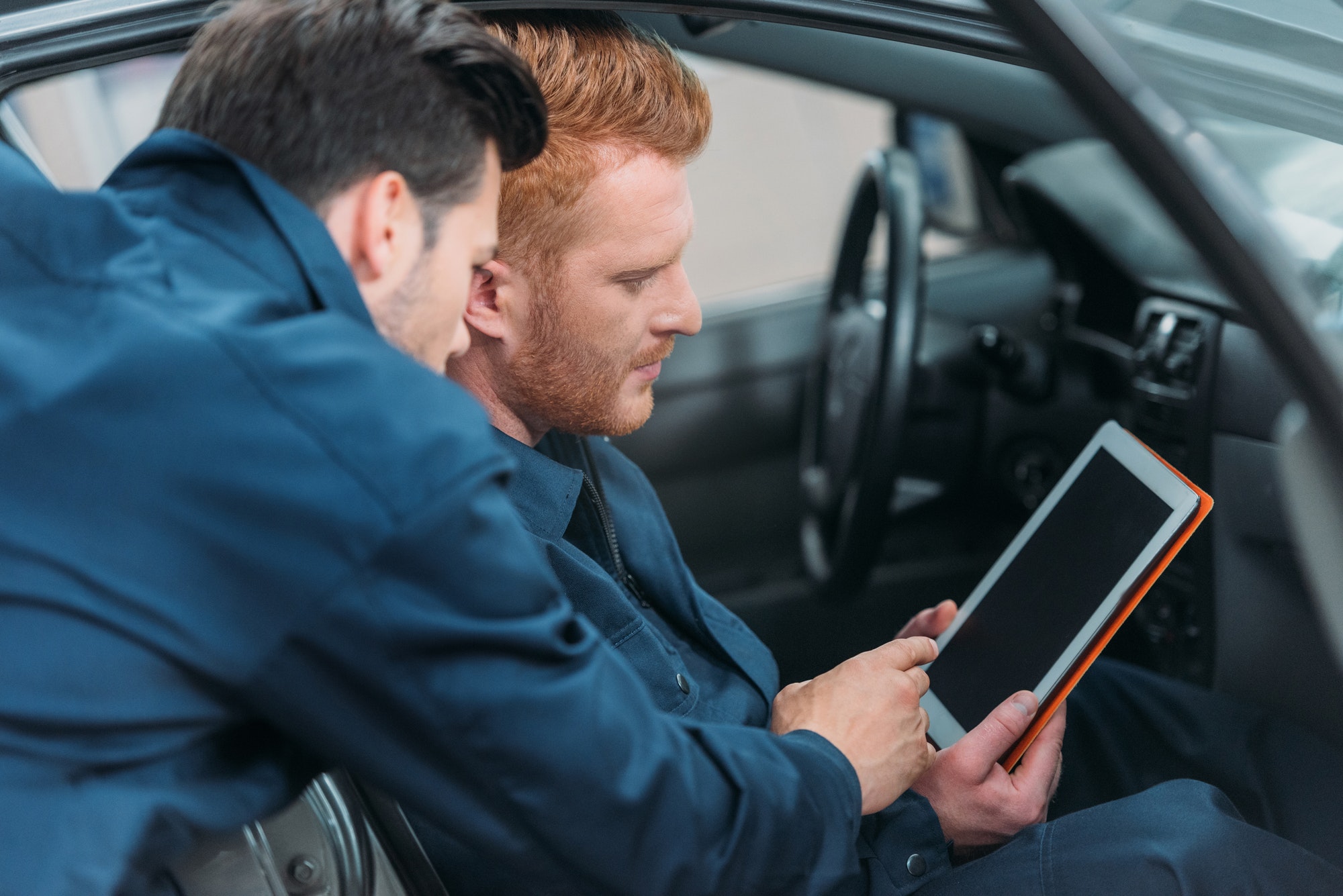 Two car mechanics sitting in a car at workshop using a digital tablet.